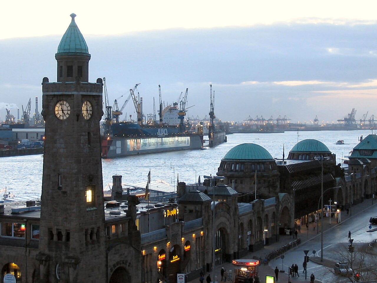 Die St. Pauli-Landungsbrücken in Hamburg mit dem Pegelturm und den Hafenkränen an der Elbe in der Abenddämmerung