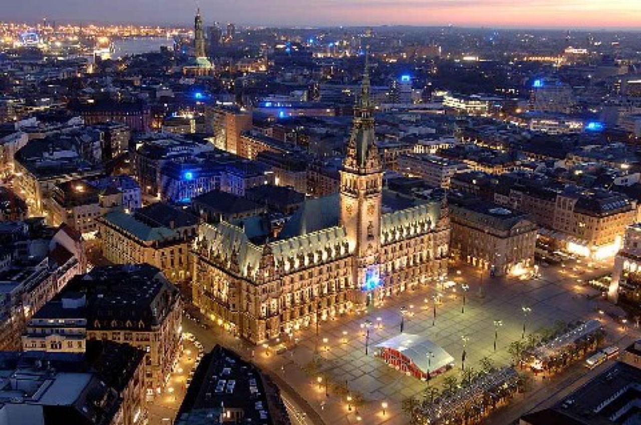 Luftaufnahme des beleuchteten Hamburger Rathauses mit Turm und Rathausmarkt in der Abenddämmerung, umgeben von der Altstadt